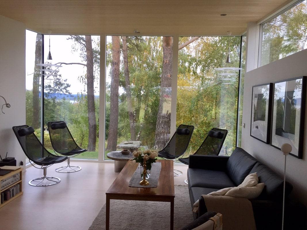 Stue med glassvegger mot furuskogen, lenestoler og sofa